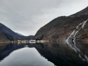 Norsk fjord med vandfald, bjerge og lille landsby omgivet af dramatisk natur og spejlblankt vand.
